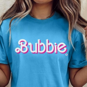 May include: A light blue t-shirt with the word "Bubbies" in a white and pink font. The shirt has short sleeves and a crew neck. The shirt is made of a soft material.