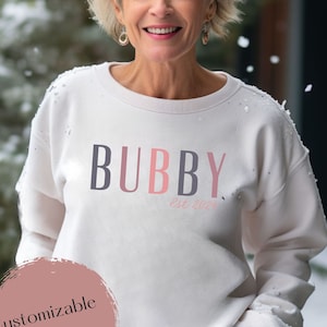 May include: White sweatshirt with the text "BUBBY Est 2024" in pink, grey, and black lettering. The year is customizable.