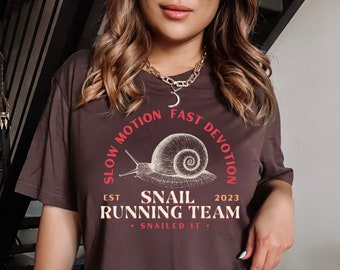 May include: A dark brown t-shirt with a white snail illustration and the text "Slow Motion Fast Devotion Est 2023 Snail Running Team - Snailed It - "