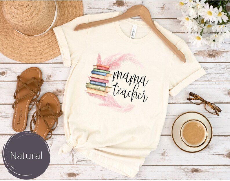 May include: A white t-shirt with a pink feather design and the text "mama teacher" in black. The shirt is laying on a wooden surface with a straw hat, sandals, and a cup of coffee.