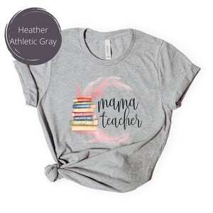 May include: Heather athletic gray t-shirt with a stack of books and the text "mama teacher" in black lettering. The books are surrounded by pink feathers.