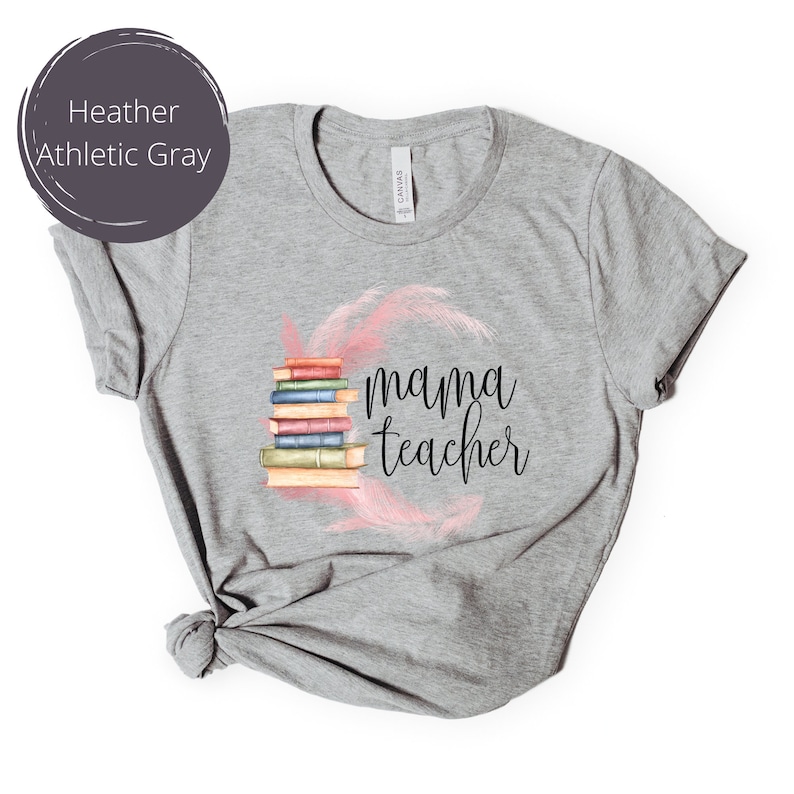 May include: Heather athletic gray t-shirt with a stack of books and the text "mama teacher" in black lettering. The books are surrounded by pink feathers.