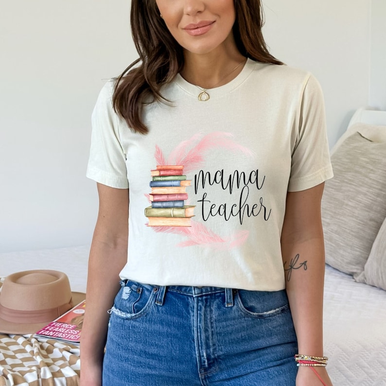 May include: A white t-shirt with a stack of books and pink feathers around them. The text "mama teacher" is written in black below the books.
