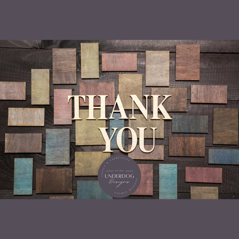 May include: A wooden sign with the words "THANK YOU" in large, block letters. The sign is surrounded by smaller wooden squares in various shades of brown, red, and blue. The sign also includes a small circle with the text "UNDERDOG Designs" in white lettering.