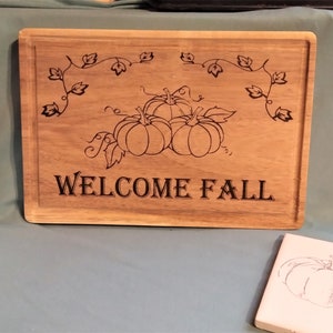 May include: A rectangular wooden sign with the words "WELCOME FALL" etched in black. The sign features a design of three pumpkins and vines. The sign is light brown in color.