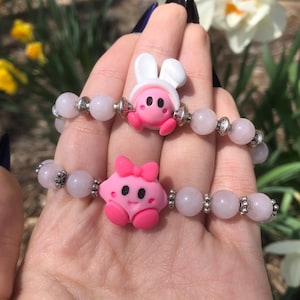 May include: Two pink and white charm bracelets with pink and white beads. One charm is a pink and white bunny with a white hat. The other charm is a pink and white character with a bow.