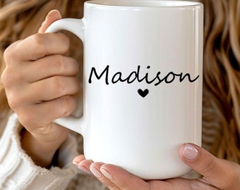 May include: A large white ceramic mug with the name "Madison" in black script and a small black heart. The mug has a large handle and is held by a person wearing a beige sweater.