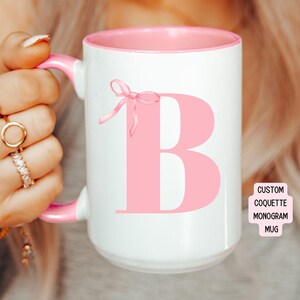 May include: White ceramic mug with a pink rim and handle. The mug features a large pink letter "B" with a pink bow on the left side. The text "CUSTOM COQUETTE MONOGRAM MUG" is printed on a white rectangle in the lower right corner.