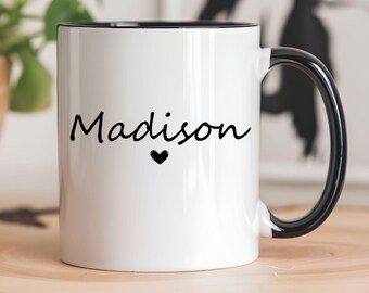 May include: White ceramic mug with a black interior, handle, and the name "Madison" in black script, with a small black heart below the name. The mug is sitting on a wooden surface.