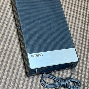 May include: A rectangular black Sony speaker with a ribbed front panel and a silver metallic base featuring the Sony logo. The device includes a thin black electrical cord with a standard audio jack connector extending from the bottom edge.