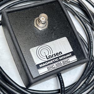 May include: A black rectangular Larsen magnetic mount with a BNC connector on top. A silver label displays the Larsen logo, the text MAGNETIC MOUNT, and BNC-MM-BNC. A long black coaxial cable with a BNC connector is attached to the device.