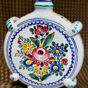 May include: A white ceramic canteen with a round body, a narrow neck, and two side handles. The front is decorated with a hand-painted floral motif featuring red, yellow, and blue flowers with green leaves, framed by a blue scalloped border.