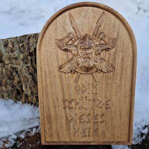 Pode incluir: Uma placa de madeira com uma imagem esculpida de Odin, o deus nórdico da sabedoria, da guerra e da morte. A placa apresenta o icônico olho único de Odin, sua longa barba e dois corvos. A inscrição diz "Odin Schütze Dieses Heim".