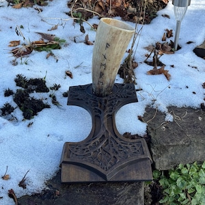 Pode incluir: Um chifre de bebida viking com runas esculpidas, exibido em um suporte de madeira em forma de martelo de Thor. O chifre é de cor clara e o suporte é marrom escuro. O fundo mostra neve, folhas e vegetação.