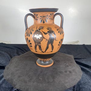 May include: A terracotta-colored amphora with black handles and a black base. The vase features black and white figures and floral designs. The figures appear to be engaged in combat. The vase is displayed on a dark gray surface.