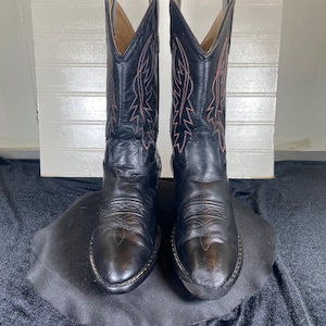 May include: A pair of black leather cowboy boots with decorative stitching in a contrasting pink color. The boots have pointed toes and a classic Western design, suitable for riding or casual wear.