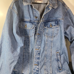 May include: A blue denim jacket with a Wrangler logo on the left chest pocket. The jacket has a button closure and two front pockets.