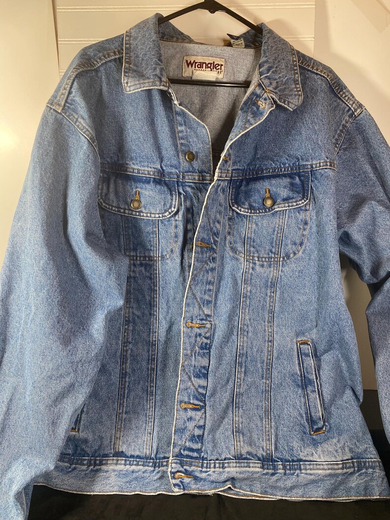 May include: A blue denim jacket with a Wrangler logo on the left chest pocket. The jacket has a button closure and two front pockets.