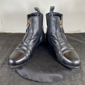May include: A pair of black leather ankle boots with a front zipper closure. The boots have a rounded toe and a small heel. The zippers have gold-colored pulls. Green and white fabric loops are at the top of the boots.