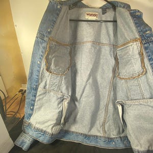 May include: A light blue denim jacket with a Wrangler label on the inside. The jacket has two pockets on the inside and is unbuttoned.
