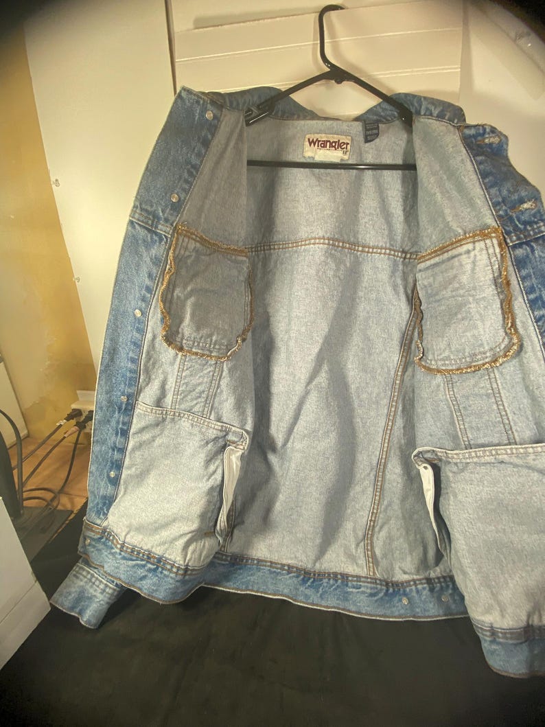 May include: A light blue denim jacket with a Wrangler label on the inside. The jacket has two pockets on the inside and is unbuttoned.