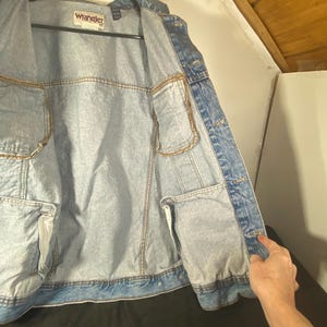 May include: A light blue denim jacket with a Wrangler label on the front. The jacket is unbuttoned and the inside lining is visible.