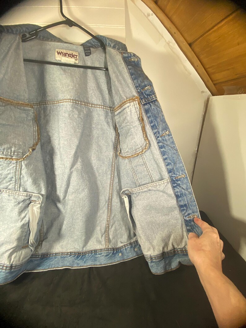 May include: A light blue denim jacket with a Wrangler label on the front. The jacket is unbuttoned and the inside lining is visible.