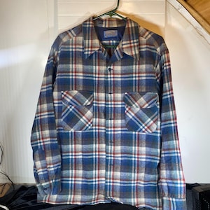 May include: A blue, red, and white plaid shirt with two chest pockets hangs on a green hanger. The shirt has a button-down front and long sleeves. The shirt is made of a soft, woven fabric.