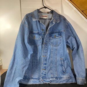 May include: A light blue denim jacket with a Wrangler label on the left chest. The jacket has a button closure, two chest pockets, and two front pockets.