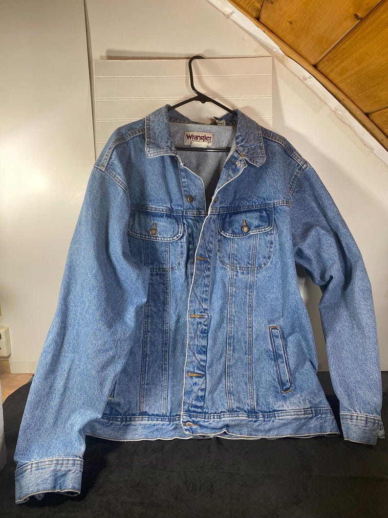 May include: A light blue denim jacket with a Wrangler label on the left chest. The jacket has a button closure, two chest pockets, and two front pockets.