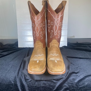 May include: A pair of brown and tan leather cowboy boots. The boots have a square toe and are decorated with white floral embroidery. The shaft of the boots is a darker brown, while the foot is a lighter tan color.