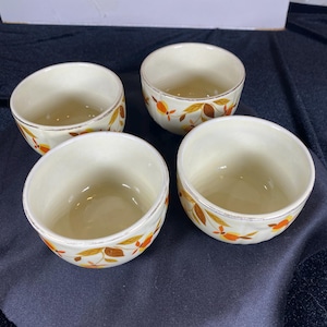 May include: Four off-white ceramic bowls with a leaf design in orange, yellow, and brown. The bowls have a rounded shape and a gold rim. The bowls are arranged on a dark surface.