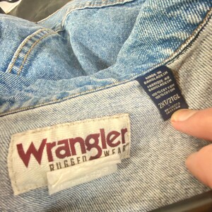 May include: A blue denim jacket with a white Wrangler Rugged Wear label and a blue care label that reads "Made in Bangladesh, Fabrique au Bangladesh, 100% Cotton, 100% Coton, 2XT/2TGL".