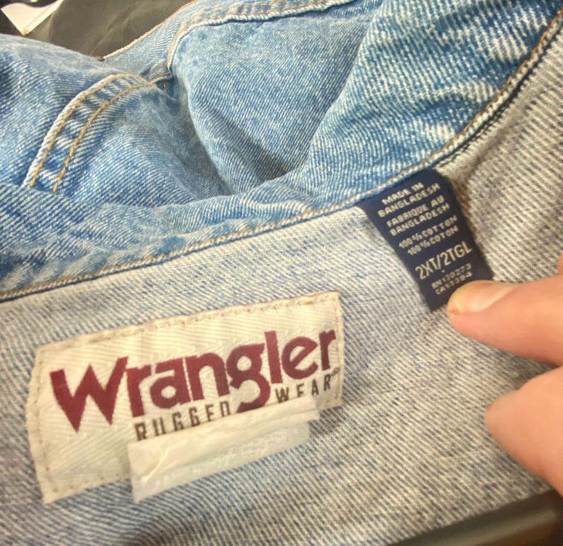 May include: A blue denim jacket with a white Wrangler Rugged Wear label and a blue care label that reads "Made in Bangladesh, Fabrique au Bangladesh, 100% Cotton, 100% Coton, 2XT/2TGL".