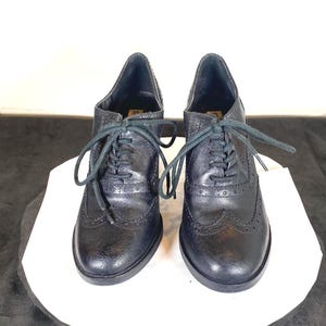May include: A pair of black leather oxford shoes with a brogue design. The shoes have laces and a low heel. The shoes are displayed on a white surface against a neutral background.