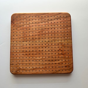 May include: A square wooden cutting board with a brown finish and a pattern of evenly spaced holes drilled throughout the surface.