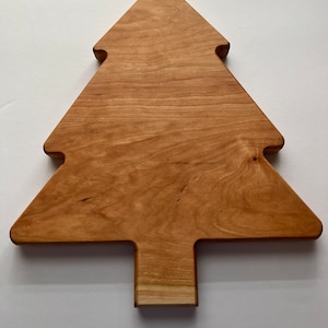 Puede incluir: Una tabla de cortar de madera con forma de árbol de Navidad. La tabla es de madera marrón claro con un acabado natural.