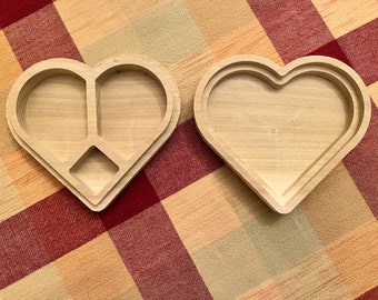Puede incluir: Dos cajas de madera sin terminar en forma de corazón. Una caja tiene una tapa con un contorno en forma de corazón y un interior dividido. La otra caja está abierta, mostrando la forma de corazón y un interior empotrado. Las cajas son de madera clara.