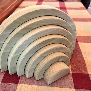 Op de afbeelding: Een houten regenboog stapelspeelgoed met zeven gebogen stukken. Het natuurlijke hout heeft zichtbare nerven en is in een halve cirkel gerangschikt. Het speelgoed staat op een rood-wit geruit tafelkleed. Een klassiek educatief speelgoed.