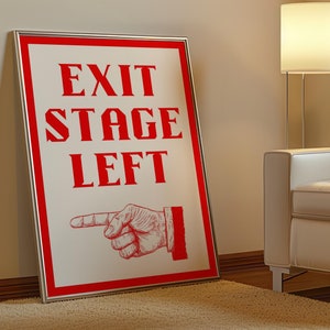 May include: A white sign with a red border and red text that reads "EXIT STAGE LEFT". A red hand with a pointing finger is pointing to the left. The sign is hanging on a white wall in a room with a white armchair and a lamp.