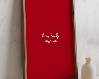 May include: A framed art print with a vibrant red background and the handwritten text "how lucky are we" in white. The frame is a light wood color. A small, cream-colored vase sits nearby.
