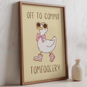 May include: Framed art print with a cartoon goose wearing sunglasses, a pink bow, and plaid boots. The text reads "OFF TO COMMIT TOMFOOLERY". The artwork is set against a light yellow background. The frame is brown.