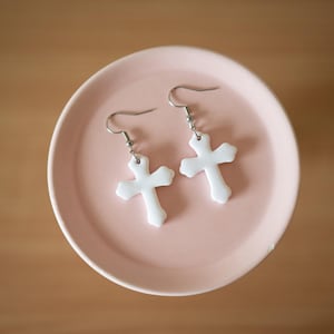 May include: A pair of white cross-shaped dangle earrings featuring silver-toned metal fishhook wires. The earrings have a smooth, opaque finish and a classic four-pointed cross design with rounded edges, suspended from simple metal hooks.
