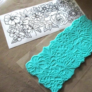 May include: A teal blue rubber stamp with a floral design. The stamp is on a clear plastic sheet.