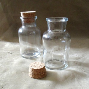 May include: Two clear glass bottles with cork stoppers. One bottle has a cork stopper in place, while the other bottle has the cork stopper off and lying on a light brown surface.