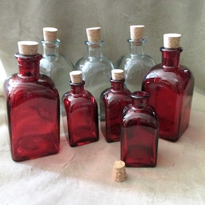 May include: A collection of eight glass bottles with cork stoppers.  The bottles are in two colors: clear and red.  The red bottles are a deep, rich red.