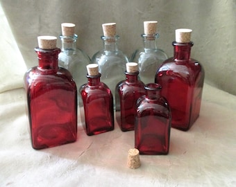 May include: A collection of eight glass bottles with cork stoppers.  The bottles are in two colors: clear and red.  The red bottles are a deep, rich red.