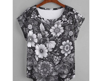 May include: A black and white floral print t-shirt with a scoop neck and short sleeves. The shirt features a detailed pattern of flowers and leaves in varying shades of gray and white, creating a monochromatic design. The shirt is hanging on a wooden hanger.