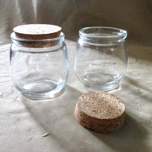 May include: Two clear glass jars with cork lids. The jars are round and have a wide mouth. The lids are brown and have a natural cork finish.