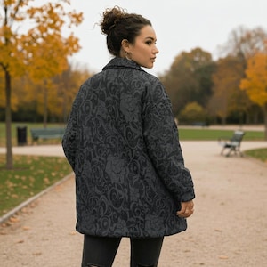 May include: A dark gray, quilted jacket with a floral pattern. The jacket has a button-up front and a collar. The person is wearing black jeans. The background is a park with trees and a walking path.